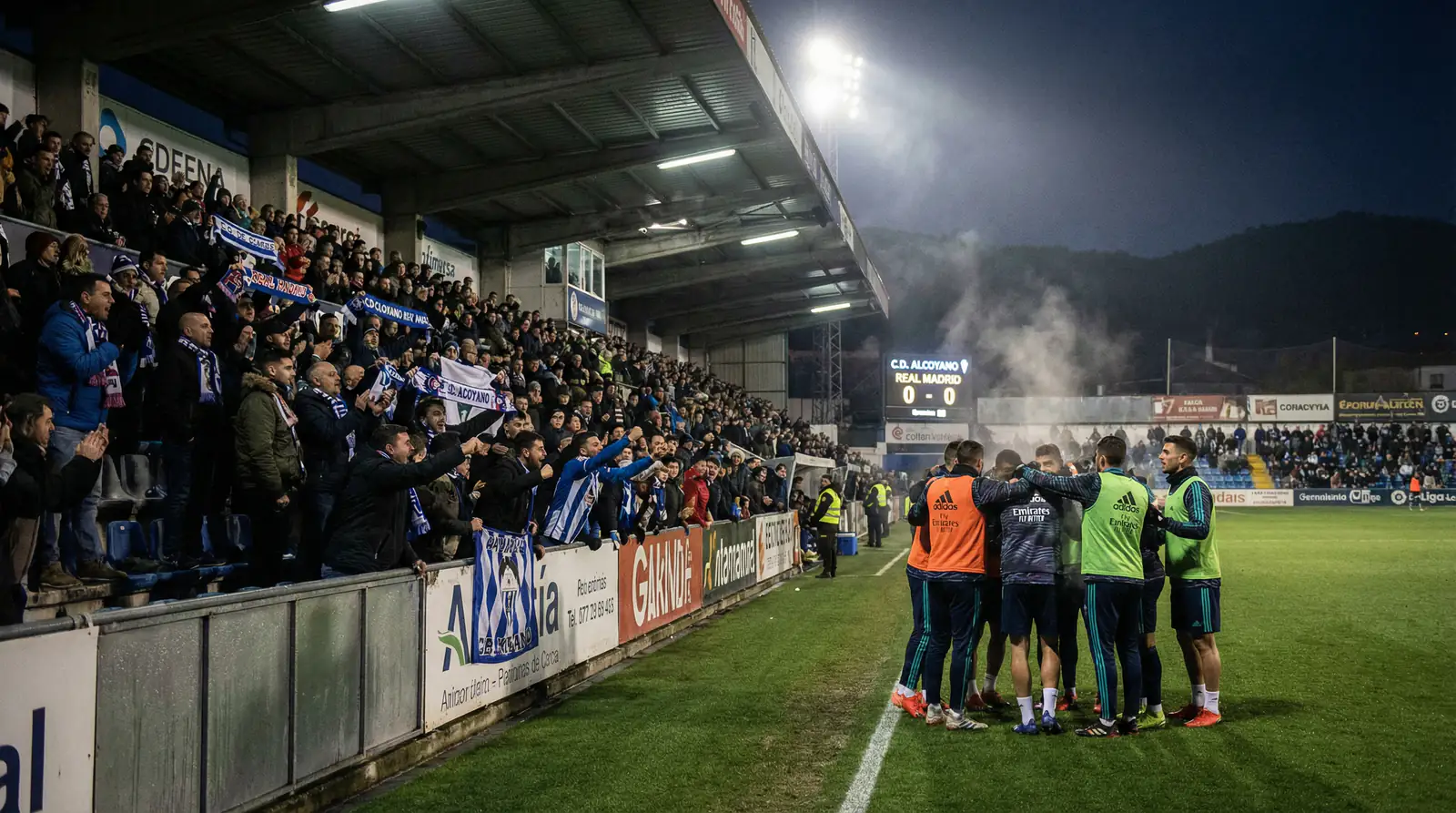 Estadio modesto de categoria inferior con cesped artificial y aficionados locales creando ambiente intimidante en noche de Copa del Rey