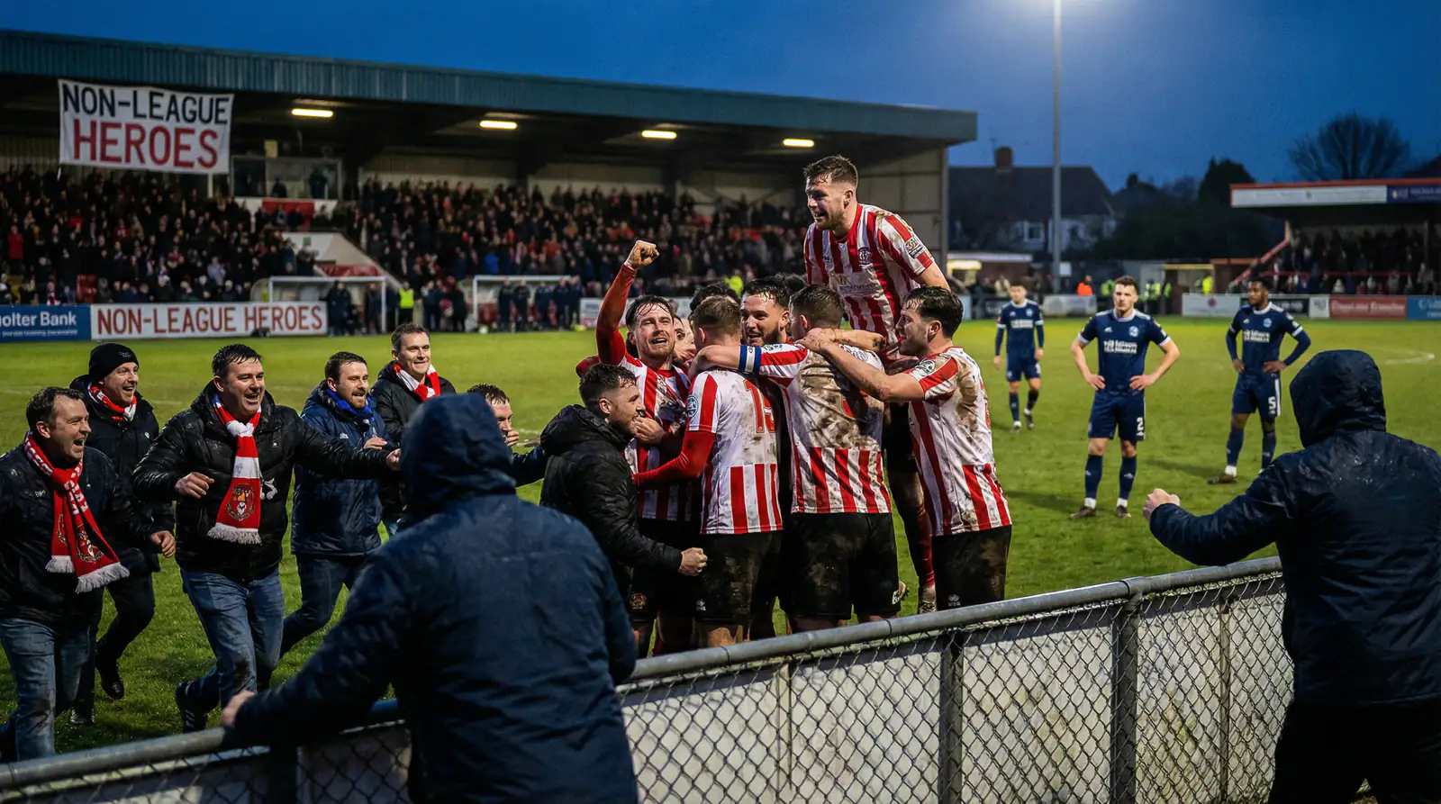 Jugadores de equipo modesto celebrando apasionadamente una victoria sorpresa de copa contra un rival de elite mientras los aficionados locales invaden el campo